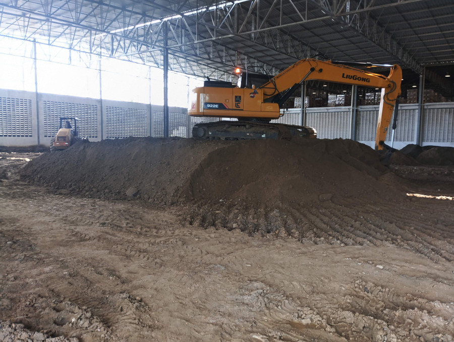 Escavadeira e rolo compactador nivelando piso dentro de galpão industrial em obra